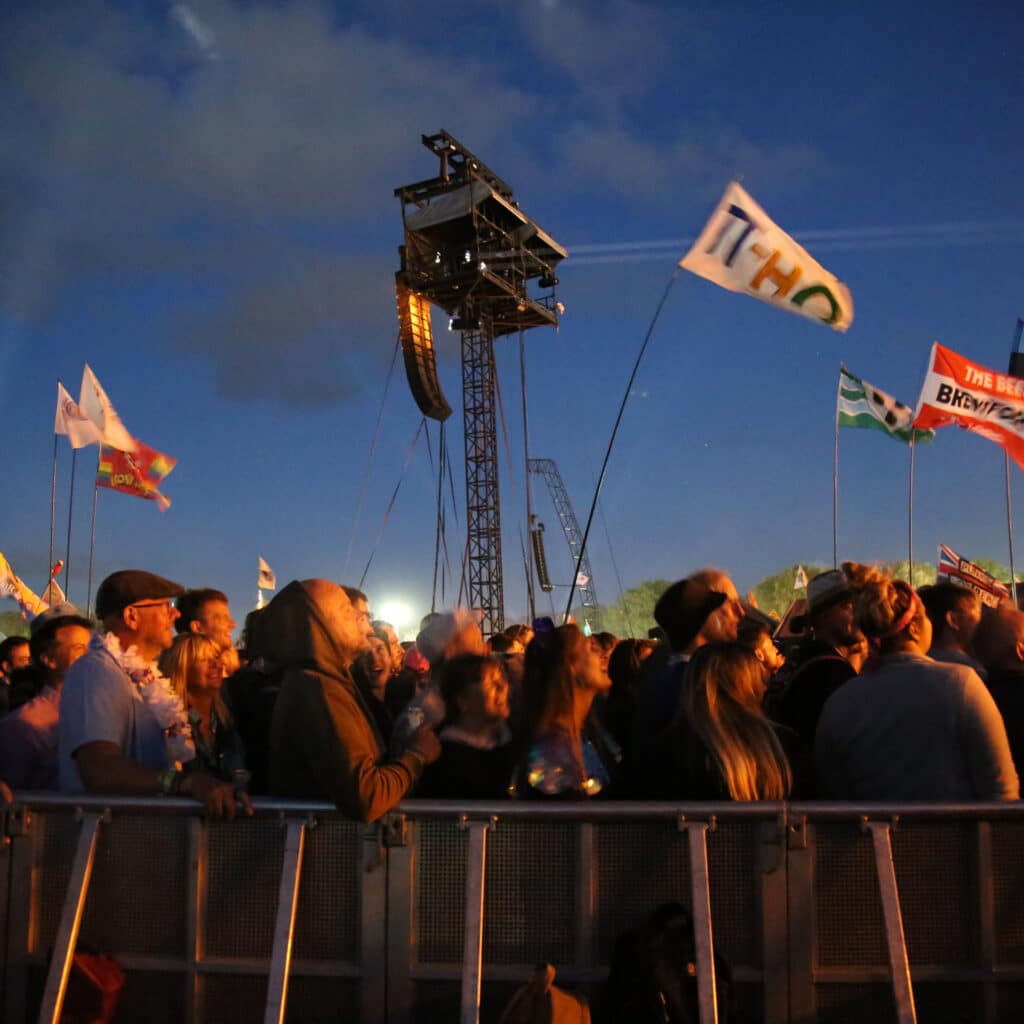 Crowd behind fence at Glastonbury at night. Faces are glowing from the yellow stage lights. Flags fly above their heads. Large audio and light tower stands in the middle