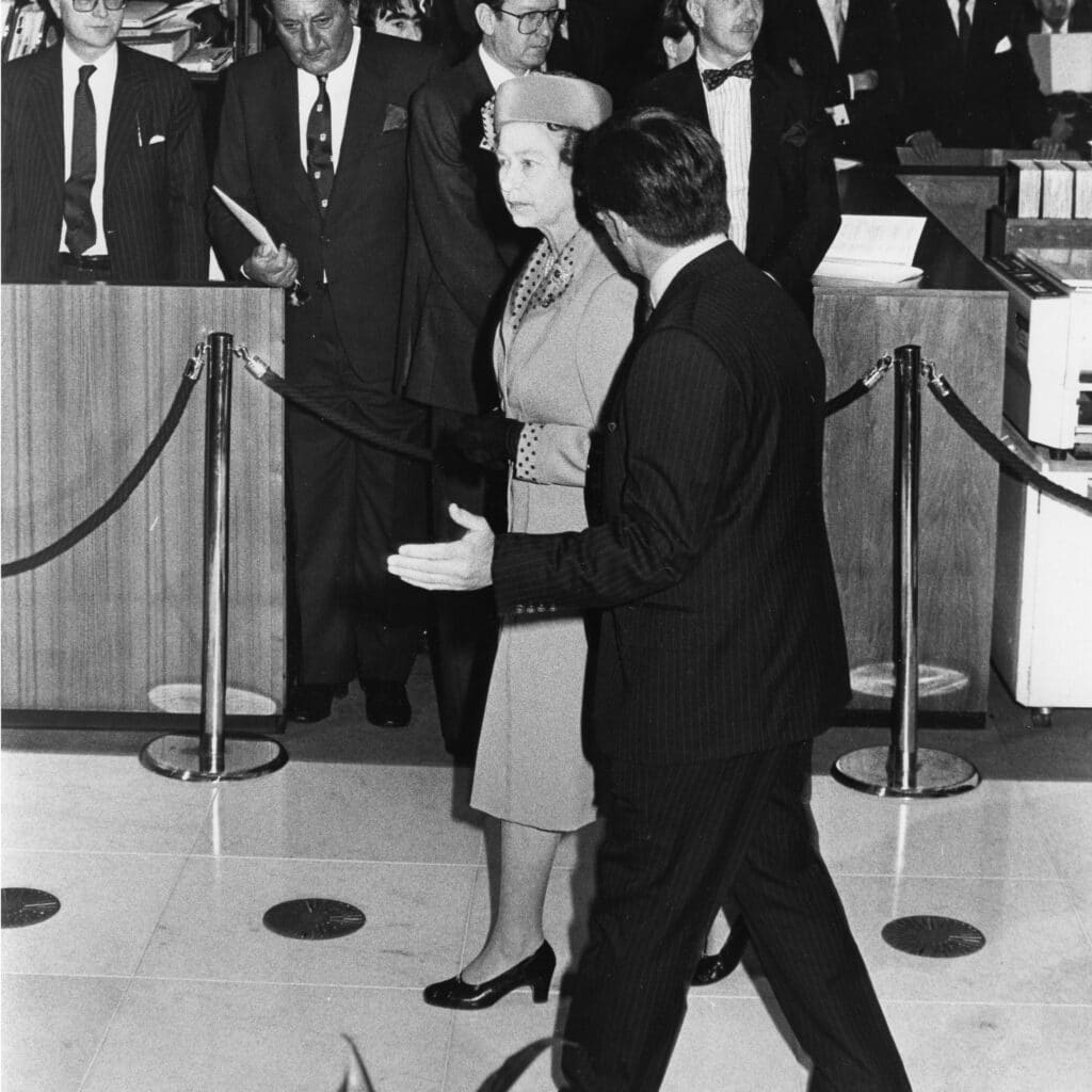 Black and white image of the late Queen Elizabeth II walking with male in pinstriped suit.